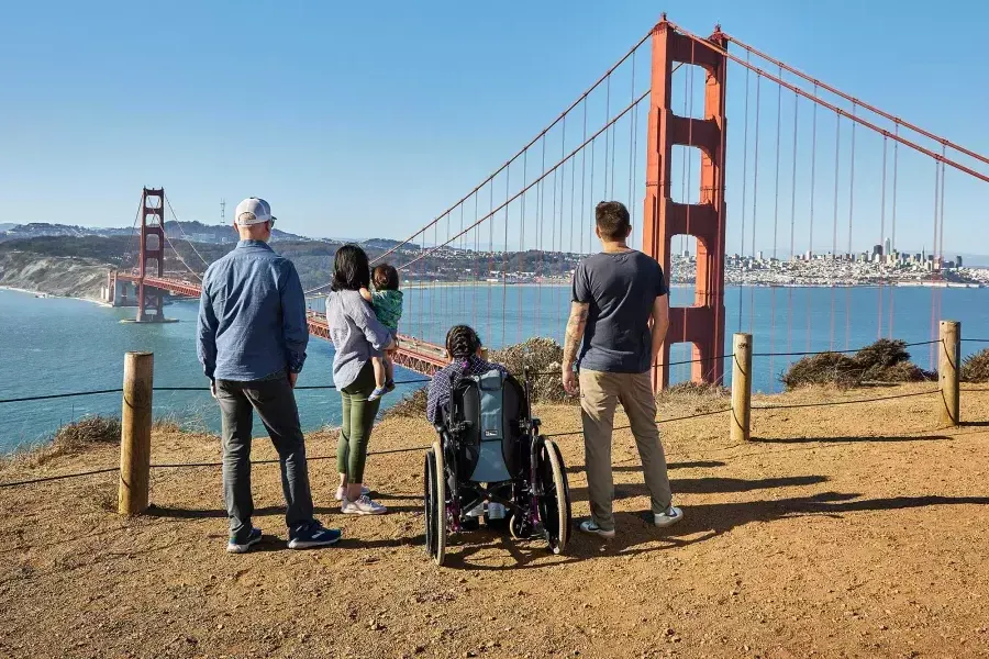 휠체어를 탄 사람 한 명을 포함한 사람들의 그룹이 마린 헤드랜드에서 Golden Gate Bridge 바라보고 있는 모습이 뒤쪽에서 보입니다.